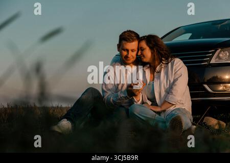 Couple holding hands sitting near car at sunset Stock Photo