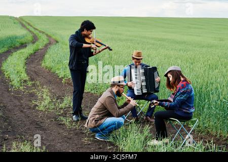 Group of young adult multiethnic musicians performing outdoors in green field, standing and sitting while playing violin, accordion, guitar and flute, focused on instruments Stock Photo