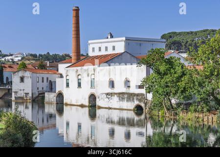A large, white industrial building with a prominent red brick chimney stands beside a calm river, its reflection visible in the tranquil water. A symb Stock Photo