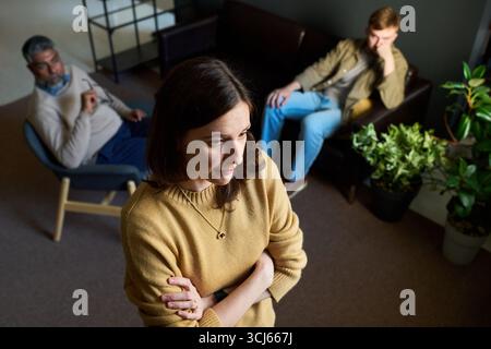 Caucasian young adult woman standing with arms crossed in foreground, middle aged Caucasian man and young adult Caucasian man sitting on chairs in background, all looking thoughtful Stock Photo