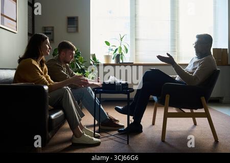 Caucasian young adult woman and Caucasian young adult man sitting on couch, talking with middle aged Indian man therapist during counseling session in modern office Stock Photo