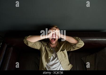 Caucasian young adult man sitting on couch covering face with hands and screaming, expressing intense frustration or stress, head tilted back, mouth wide open, eyes closed Stock Photo