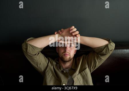 Portrait of young adult Caucasian man reclining on dark sofa, gazing directly into camera with neutral expression, resting arms above head, hands interlaced on forehead Stock Photo