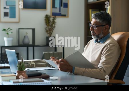Middle aged Indian man sitting at desk reviewing documents while working on laptop in modern office, wearing eyeglasses and concentrating on paperwork, indoor business setting Stock Photo