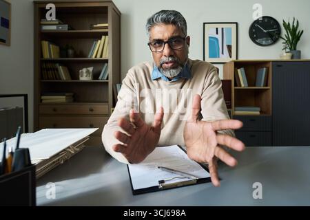 Portrait of middle aged South Asian man gesturing with hands while sitting at desk with documents and clipboard, looking into camera during online business meeting or consultation Stock Photo