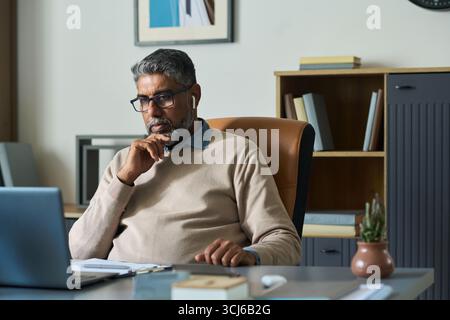 Middle aged South Asian man sitting at desk working on laptop, wearing eyeglasses and wireless earphones, resting chin on hand, appearing focused in modern office setting Stock Photo