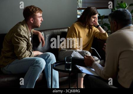 Caucasian young adult man and Caucasian young adult woman sitting on sofa talking with middle aged man during counseling session, woman appearing thoughtful, man listening attentively Stock Photo