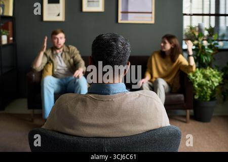 Back view of middle aged multiethnic man sitting and listening to young Caucasian man and young Caucasian woman having conversation in office setting, during counseling session Stock Photo