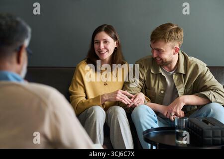 Caucasian young adult woman and Caucasian young adult man sitting on couch holding hands, smiling and talking with middle aged Black man during counseling session Stock Photo