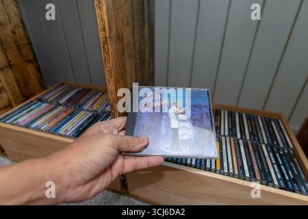 Close-up of hand holding a retro music CD labeled "Slow Dancing" in front of wooden shelf full of colorful CDs. Home music collection, nostalgia, and Stock Photo
