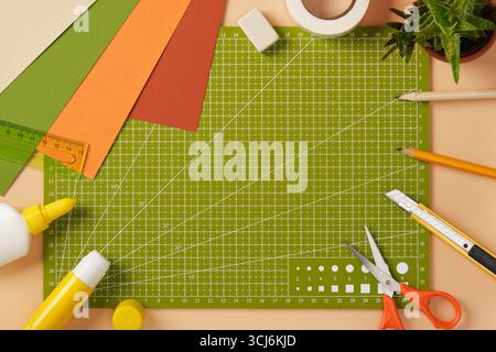 Creative mess on the paper crafting table. Colored sheets of paper, glue, pencils, eraser, scissors, and knife on a cutting mat. Stock Photo
