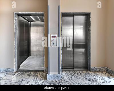 Two stretcher elevators, one lift enclosed with stainless steel doors and the other open and empty on the floor with white marble flooring in the hosp Stock Photo