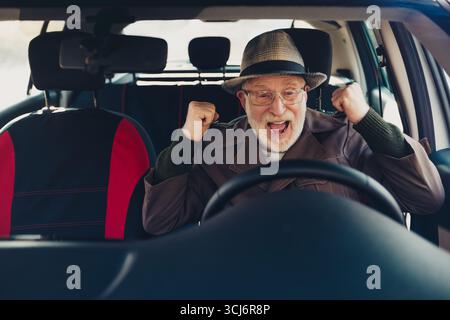 Happy elderly man cheering with excitement while sitting in a car on a sunny day, enjoying the new driving experience Stock Photo