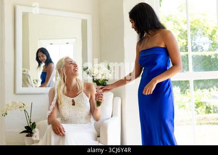 Bride holding bouquet, smiling with bridesmaid in blue dress by large window Stock Photo
