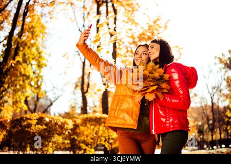 Two friends taking a selfie in an autumn park surrounded by golden foliage, enjoying the season in vibrant casual wear Stock Photo