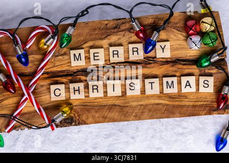 Merry Christmas spelled out using wooden letter tiles on a rustic wooden background; surrounded by colorful Christmas lights; candy canes; and festive Stock Photo