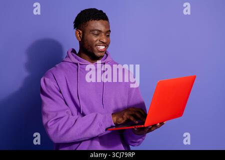 Young man holding a laptop and smiling, dressed in purple sportswear, standing against a vibrant purple background Stock Photo