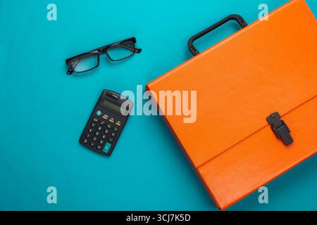 Orange paper case, calculator, glasses on a blue background. Top view Stock Photo