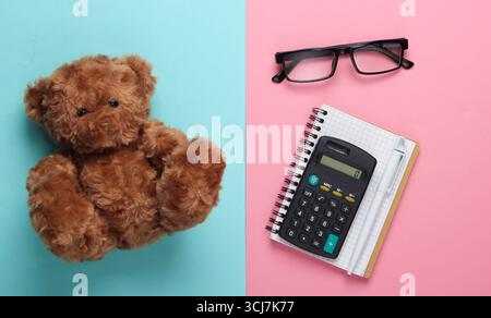 flat lay composition of a teddy bear, notebook with pen, calculator, glasses on a blue-pink pastel background. Top view Stock Photo