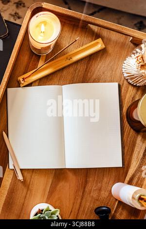 Cozy workspace for journaling or meditation with an open notebook on a wooden tray surrounded by scented candles and incense Stock Photo
