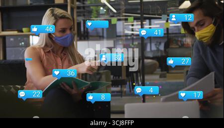 Two masked colleagues discussing binder in open-plan office, with floating icons and sticky notes Stock Photo