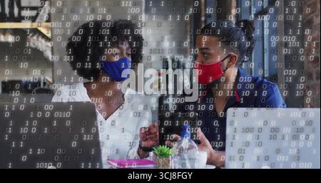Discussing project two masked colleagues using laptops in cafe, with succulent and binary overlay Stock Photo