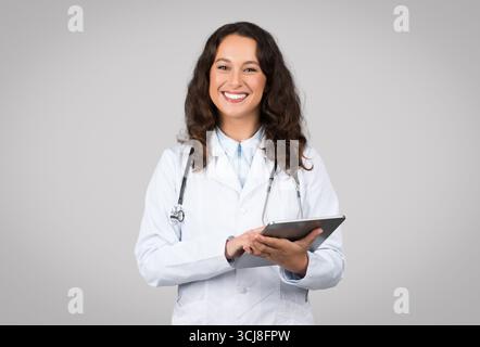 Positive woman in medical workwear doctor using digital tablet isolated on grey background, chatting with patient Stock Photo