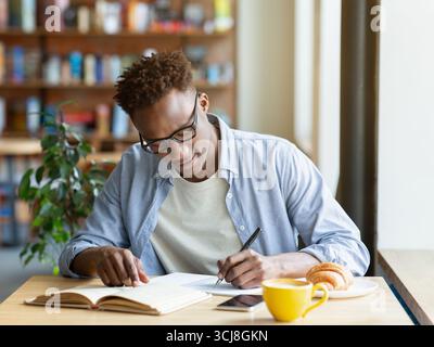 Hardworking black man writing down info from textbook, getting ready for test at cafe Stock Photo