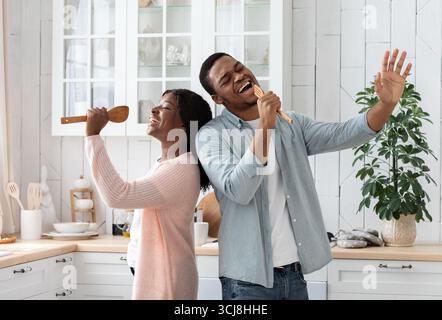 Domestic Concert. Playful African Couple Having Fun In Kitchen, Singing And Dancing Stock Photo