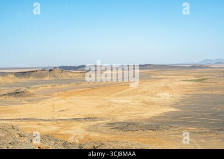 Vast Sahara desert plain in Morocco with golden sand patches dark rocky hills and distant horizon under clear blue sky creating wide panoramic view Stock Photo