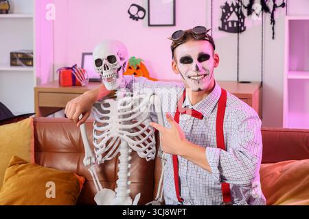 Young happy man dressed for Halloween pointing at skeleton on sofa in decorated living room Stock Photo