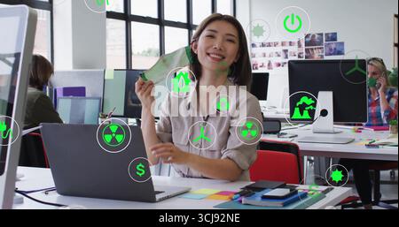 Smiling Asian graphic designer holding green concept card at open-plan office desk, with monitors Stock Photo