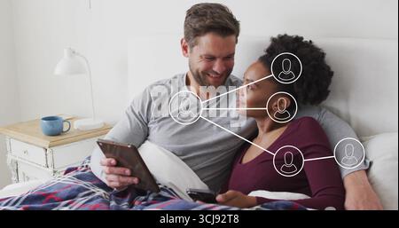 Using tablet and smartphone, couple wearing long-sleeve tops lounging on bed, with plaid blanket Stock Photo