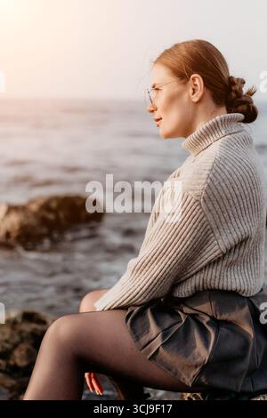 Woman ocean sitting, thoughtful young adult female on rocks by the sea at sunset Stock Photo