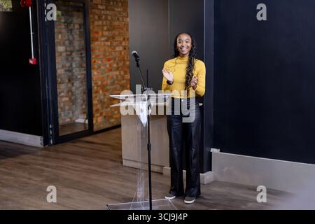 African American woman in mustard top clapping behind clear lectern with microphone in event space Stock Photo