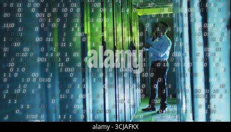 IT technician analyzing server rack in data center corridor, with tablet and floating binary code Stock Photo