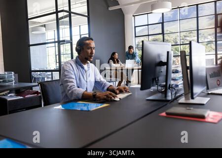 In office, Asian man using computer with headset, colleagues collaborating nearby Stock Photo
