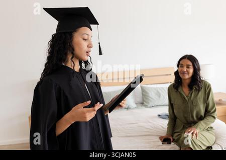 Asian graduate in cap and gown reading speech at home with proud mother watching Stock Photo