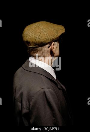 An older man wearing a tweed cap, glasses, and a dark coat, seen from the back, against a black background. Stock Photo