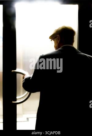An older man wearing a hat and glasses is opening a door. Stock Photo