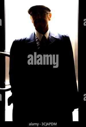 An older man wearing a hat, glasses, and a suit stands in a doorway, silhouetted against the bright light behind him. Stock Photo