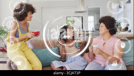 Chatting three friends holding mugs around sofa in bright living room, with digital interface theme Stock Photo