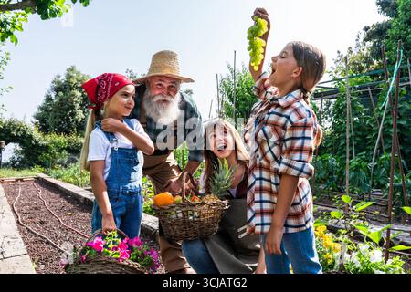 Happy farmers family with grandfather, mother and daughters harvesting organic grapes, pineapple and oranges in wicker basket working together in agri Stock Photo