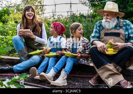 Cheerful family farmers enjoying a healthy lunch of sandwiches together in their vegetable garden on a bio farm, promoting a sustainable and healthy l Stock Photo