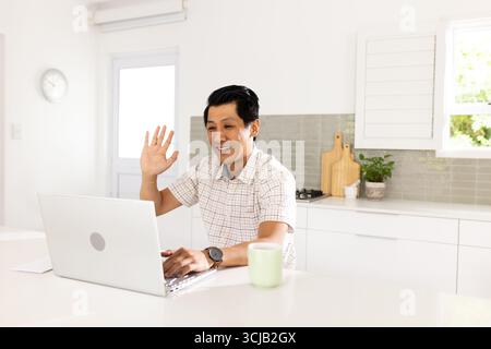 Asian man video chatting on laptop in kitchen, smiling and waving hello Stock Photo