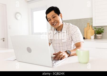 Smiling man using laptop at home, enjoying coffee in bright kitchen Stock Photo
