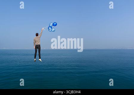 A boy flies over the sea, carried by balloons. The image symbolizes freedom, dreams, escape, and the start of new adventures with ample space for text Stock Photo