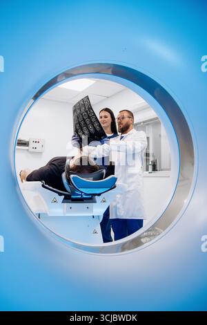 Medical professionals in white lab coats are reviewing CT scan images while assisting a patient on an examination table in a modern medical facility, Stock Photo