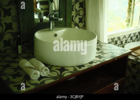A round white ceramic washbasin with a faucet, mirror, and rolled towels in a patterned tile bathroom Stock Photo