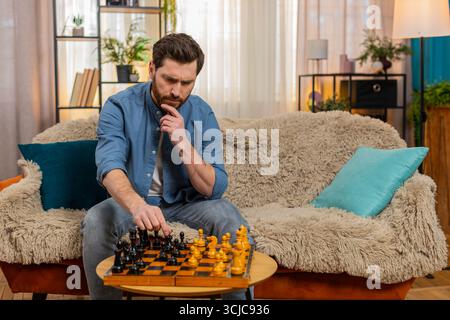 Young man sitting at home on sofa, playing chess alone, making a serious move with deep concentration. Caucasian guy strategizing, feeling thoughtful and focused, symbolizing dual personality indoors Stock Photo